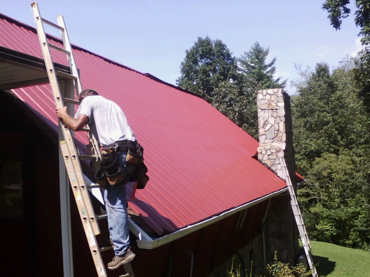 Roofer on a ladder beside a red metal roof and stone chimney — Soffit & Fascia Repair by Roofing Brothers in Goldendale